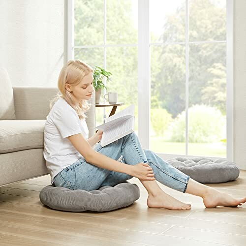 Woman sitting on a floor cushion reading a book near a window.